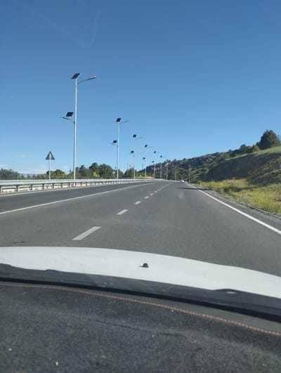 A forward-facing, eye-level view captures a highway under a clear blue sky. Solar-powered streetlights line the left side of the road, spaced evenly apart. The highway is asphalt with white dashed lane markers. To the right of the highway, a grassy, hilly terrain with sparse trees and rocky outcrops rises. The foreground shows the dashboard and windshield of a vehicle, indicating the image was taken from inside a car. The location is the M37 highway approaching the city of Myk, Uzbekistan, characterized by its arid landscape and solar-powered infrastructure.