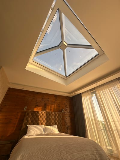 The image shows a bedroom with a large, modern skylight in the ceiling. The skylight has a geometric design with multiple glass panels. The room features a queen-size bed with a tufted headboard and white pillows. The walls are paneled with dark wood, and there are sheer curtains covering a window to the right, allowing sunlight to stream into the room. The overall impression is one of modern luxury with a touch of rustic charm. The image does not provide any specific information about the location being Brca, Montenegro.