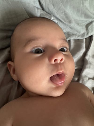 A close-up, low-angle shot captures an infant's face and upper torso. The baby has dark eyes, a small nose with flared nostrils, and an open mouth with its tongue slightly protruding, glistening with moisture. The skin tone is warm. The baby is lying on a wrinkled, light grey fabric, likely bedding. The background is entirely composed of this fabric, suggesting an indoor setting. The image appears to be taken at close range, with the baby's face filling most of the frame. There is no discernible information in the image that would indicate a location in Oldham, United Kingdom.