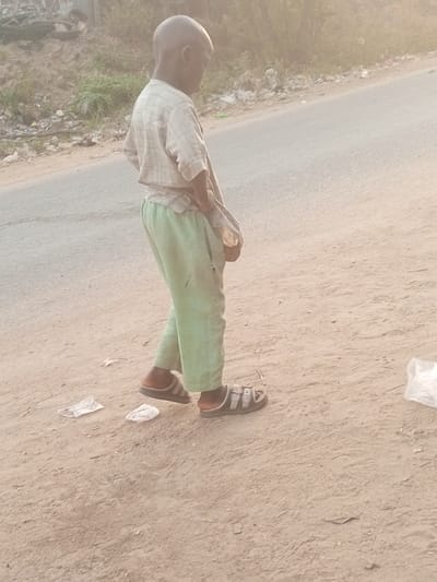 A young child, viewed from behind and slightly to the right, walks on a dusty path adjacent to a paved road. The child has short, dark hair and is wearing a light-colored, possibly striped, short-sleeved shirt and loose-fitting, light green trousers that appear somewhat worn. On their feet, the child wears two different styles of dark sandals: a slip-on sandal on the left foot and a double-strap sandal on the right.
The child's head is slightly bowed, suggesting they are looking downwards or thoughtfully ahead. One hand is positioned near the hip, possibly holding or adjusting something. The ground is dry, light brown earth or gravel, with a few small pieces of white debris scattered near the child's feet and a clear plastic bag visible on the far right. Beyond the paved road in the background, there are hints of vegetation and more scattered debris.
The lighting in the scene is soft and warm, characteristic of early morning or late afternoon, with a gentle, somewhat hazy glow that minimizes harsh shadows. The overall atmosphere suggests an outdoor, possibly semi-urban or rural setting. No text is visible in the image. The scene depicts an ordinary moment in the provided location context of Ebiya, Nigeria.
