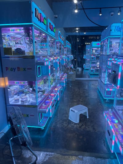 This image captures a row of brightly lit arcade claw machines in what appears to be an entertainment venue or an arcade. The machines are adorned with vibrant neon blue and pink lights, creating a futuristic and playful atmosphere. Many of the machines display the text "Play Box mini" and what seems to be Korean characters, suggesting a local establishment.
Inside the transparent compartments of the machines, various prizes, including small boxes, toys, and plush items, are visible. The floor is a dark, slightly worn concrete, and in the foreground, a metal oscillating fan is positioned, possibly to help with air circulation. A white plastic stool is also present, suggesting a waiting area or a place for patrons to rest.
The scene is predominantly lit by the artificial glow of the arcade machines, giving it a slightly surreal and eye-catching appearance. The overall impression is one of an active entertainment space, likely popular among younger audiences or those seeking amusement. The presence of text in Korean indicates this location is in South Korea, specifically noted as Cheongju-si. No people are clearly visible in the frame, but the setup implies ongoing or recent activity. The time of day is difficult to ascertain due to the artificial lighting, but the overall ambiance suggests an evening or indoor setting.