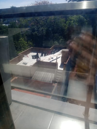 This image, taken from an elevated position such as an upper floor window, shows a building in Lomas de Zamora, Argentina. The view is partially obscured by reflections on the glass.
The building appears to be a multi-story structure with a light-colored facade and a corrugated metal roof. Laundry lines are visible on the roof, with clothes hanging to dry. The building has a cylindrical structure on its roof.
Adjacent to the building is a dense area of trees and foliage. The sky is partially visible in the upper left corner. The immediate foreground consists of the window frame. The angle of the shot suggests a downward-looking perspective.