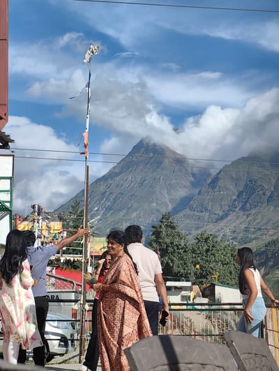 In the city of Yamling, India, a group of people are gathered on a rooftop or balcony with a scenic mountain backdrop. In the foreground, a woman in a patterned sari smiles while holding a small plant. To her left, a man in a blue shirt gestures with his hand, seemingly conversing with a woman in a floral dress and white pants. Another man, partially visible, stands with a phone in his hand. In the background, a tattered flag flies from a pole, and imposing, cloudy mountains dominate the horizon. The atmosphere appears casual and social, with the natural landscape providing a striking setting.