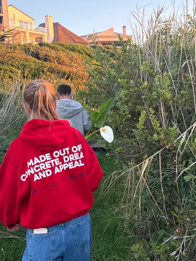 The image depicts an outdoor scene during what appears to be late afternoon or early evening, characterized by soft, warm light. In the foreground, viewed from behind, is a person with blonde hair, wearing a red hooded sweatshirt and blue jeans. White text on the back of the hoodie reads: "MADE OUT OF CONCRETE, DREA" (likely part of "DREAM") "AND APPEAL," with smaller text below indicating "ork," "City of Sky," and "est. 1975."
Slightly further back, another individual with dark hair, also seen from behind, wears a grey hoodie. Both people appear to be standing or casually walking amidst dense green vegetation, which includes tall grasses and various bushes. A single, striking white calla lily with a prominent yellow spadix stands out in the mid-ground.
In the background, modern buildings with warm-toned facades and balconies are visible through and above the foliage, suggesting a residential area adjacent to this natural setting. The sky is clear and light blue. The overall scene conveys a peaceful moment of observation or casual outdoor activity within a natural environment, likely in the Overstrand Local Municipality, South Africa, given the mix of vegetation and coastal-style architecture.