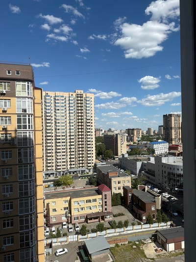 The image captures a cityscape on a sunny day, with a bright blue sky dotted with white cumulus clouds. The view is from a high vantage point, looking down onto a densely built urban area. In the foreground and to the left, a modern multi-story apartment building with a beige brick facade and numerous balconies with white-framed windows dominates the scene. To the right of this building is an even taller, slimmer apartment building with a yellow facade, densely packed with small windows.
Below these residential towers, a complex network of lower-rise buildings, likely commercial or administrative, stretches across the frame. These buildings have various architectural styles and colors, including beige, yellow, grey, and some with blue roofs. Several streets and parking lots are visible, with numerous cars parked and a few in motion. Some of the cars are white, and others are dark. Small trees and patches of greenery are interspersed among the buildings, particularly around the lower structures and in courtyard areas. A fence with a yellow top rail runs along a street. Further in the background, a dense collection of taller buildings, characteristic of a larger city, forms the horizon. The overall impression is that of a developed urban environment under clear weather conditions.
This scene is consistent with an urban setting in Novoaleksandrovka, Russia, likely a residential or mixed-use district with modern construction alongside older buildings.