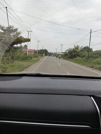 This image, taken from inside a vehicle, captures a view of a two-lane asphalt road in Tanjongkeumala, Indonesia. The road is bordered by lush green vegetation, including trees and tall grasses. Overhead, a complex network of power lines crisscrosses the sky, supported by utility poles. On the left side of the road, a red-roofed building is visible amongst the greenery. Further down the road, two motorcycles with riders are traveling in the same direction. The sky is overcast, suggesting a cloudy day. The foreground of the image shows the interior of the vehicle, with the dashboard and windshield visible.