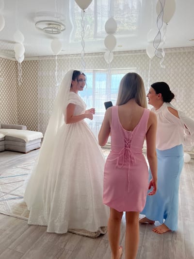 In a room adorned with white balloons suspended from the ceiling by ribbons, a bride in a sparkling white wedding dress stands to the left. She is looking towards two women on the right. The woman in the middle wears a pink dress with a lace-up back, and the woman to her right is in a white blouse with cut-out sleeves and a light blue skirt. The room has patterned wallpaper, sheer curtains, and a modern light fixture on the ceiling. A sectional sofa is visible in the background. The flooring is light-colored wood laminate, with a patterned rug in the foreground. The scene appears to be a pre-wedding preparation or celebration. The location is identified as Astrakhanka, Kazakhstan.