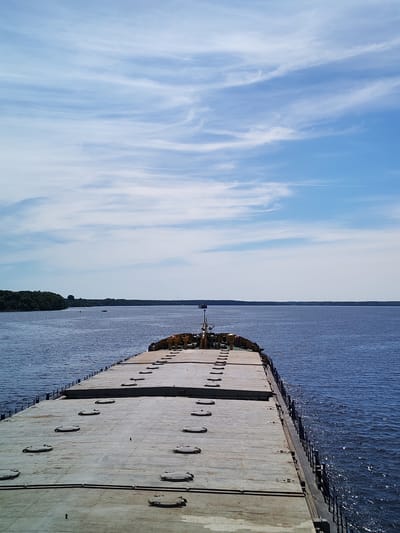 The image depicts the forward deck of a large, flat-topped cargo vessel, likely a barge or bulk carrier, underway on a wide body of water. The deck, a light grey concrete or metal surface, features numerous evenly spaced, oval-shaped hatch covers. A series of dark vertical stanchions line the port side of the deck, forming a simple railing, with a similar structure visible on the starboard side. Towards the far end of the vessel, a small superstructure or pilot house is visible, with yellow and green elements.
The vessel navigates a vast, calm expanse of dark blue water, reflecting scattered sunlight. Distant landmasses, heavily forested with green trees, line both the port and starboard horizons, suggesting a wide river or a large lake. A small red boat and another dark, indistinct vessel are visible in the distance on the port side, closer to the tree line. On the starboard side, further down the waterway, a dark, vertical structure, possibly a mast or tower, rises above the distant tree line.
The sky above is a clear, light blue with delicate, wispy white cirrus clouds stretching across the frame, indicating fair weather. A faint, linear contrail from an aircraft is visible high in the sky towards the upper right. The scene is set in the city of Priluki, Russia, on this expansive waterway.