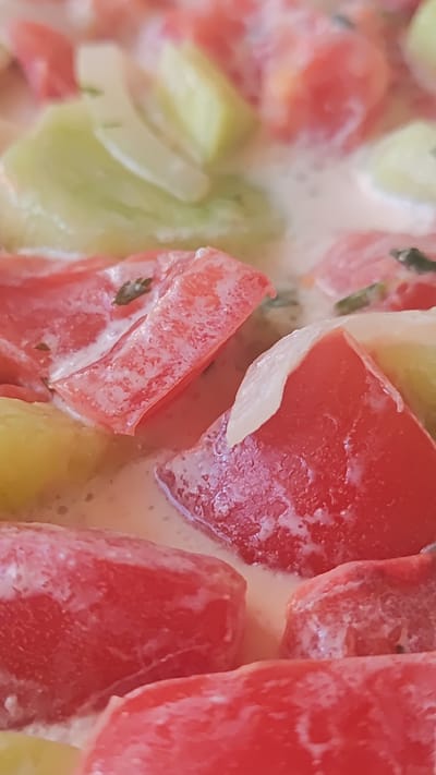 A close-up, vertical shot showcases a dish of chopped tomatoes and what appear to be pieces of cucumber or zucchini, simmering in a creamy white sauce. The ingredients are cut into roughly cubic or rectangular shapes, with the tomatoes being a vibrant red and the other vegetable a pale green. The sauce coats the pieces unevenly, creating a textured, slightly blurred appearance. Some small green herbs are visible sprinkled throughout the dish. The overall impression is of a home-cooked meal, possibly a stew or a creamy vegetable side dish. There is no discernible information about the city of Dagomys, Russia, in the provided image.