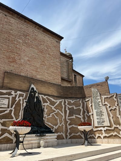 This is a photograph of a war memorial located in the city of Barbara, Italy. The memorial is made of brick and features a bronze sculpture of figures, possibly a family or group of people, in distress. The sculpture is placed on a tiered stone base in front of a brick wall decorated with a white geometric pattern. To the left of the sculpture, a marble plaque is mounted on the wall, bearing an inscription in Italian. Two large white vases filled with red flowers are positioned on either side of the sculpture, serving as planters. To the right of the sculpture, another marble plaque is affixed to the wall, also inscribed with Italian text. The memorial is situated outdoors, with a blue sky and white clouds visible in the background, as well as the side of a brick building. The scene appears to be a public space, possibly a piazza or square, with stone steps leading up to the memorial.
