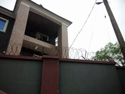 This image depicts a residential building with a dark facade and a section of brickwork on its upper floors, located in Nanka, Nigeria. A balcony is visible on the second story, featuring a black metal railing and a satellite dish. The property is enclosed by a dark green wall topped with barbed wire, suggesting enhanced security. To the right, a tall, weathered utility pole with a street lamp and hanging wires stands prominently against a pale, overcast sky. Lush green trees are visible behind the wall and barbed wire. The scene appears to be a snapshot of a typical residential area in Nanka, with elements of both modern construction and functional infrastructure.