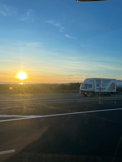 The image depicts a highway scene at sunset or sunrise near La Palme, France, overlooking the Etang de La Palme (La Palme lagoon). The sky occupies the upper two-thirds, displaying a gradient from deep blue at the top to vibrant orange and yellow along the horizon, with sparse cirrus clouds and a faint contrail visible. The sun, a brilliant white-yellow disk, is low on the left horizon, creating significant lens flare and reflecting brightly off a distant body of water. In the mid-ground, a multi-lane asphalt highway stretches horizontally, featuring a metal guardrail. A white Mercedes-Benz truck with a box body and an attached tanker trailer travels rightward on the highway. The truck's box is branded with "TDS Déménagements & Transferts 66" in blue and orange lettering, alongside the phone number "07 69 04 83 05". The landscape beyond the highway to the left and center consists of a flat expanse with a prominent body of water reflecting sunlight, and distant low-lying vegetation. The foreground exhibits dark, indistinct reflections consistent with a view from a moving vehicle. No individuals are visibly identifiable.