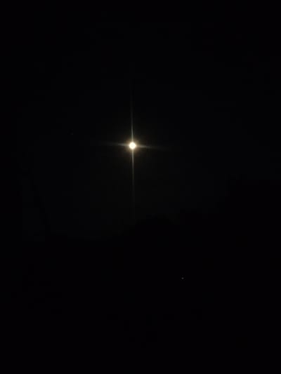 A very dark, low-resolution nighttime photograph shows a bright, star-like light source with diffraction spikes in the center of the frame. To the left of this central light source, a much fainter, smaller point of light is visible. The very bottom of the image shows a dark, indistinct horizontal shape that might be a silhouette of land or a low structure. It is not possible to determine the location within Kostiantynivka, Ukraine from the visual information provided.