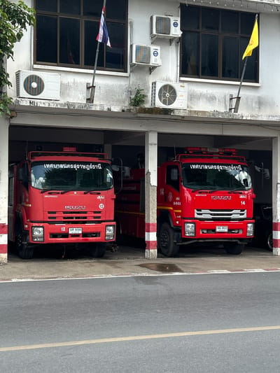 The image depicts the exterior of a fire station located in Surat Thani City Municipality, Thailand. Two red fire trucks are parked under a covered bay. The fire trucks are Isuzu models, with Thai text visible on the windshields and license plates. The truck on the left has the license plate "พร 8086" and the number "12" on its grille. The truck on the right has the license plate "พร 8622" and the number "14" on its grille. Above the bay, an outdoor area of the building is visible, featuring windows, air conditioning units, and two flags. One flag is a Thai flag, and the other is a yellow flag. The building appears to be constructed of concrete. The scene is viewed from across a paved road.