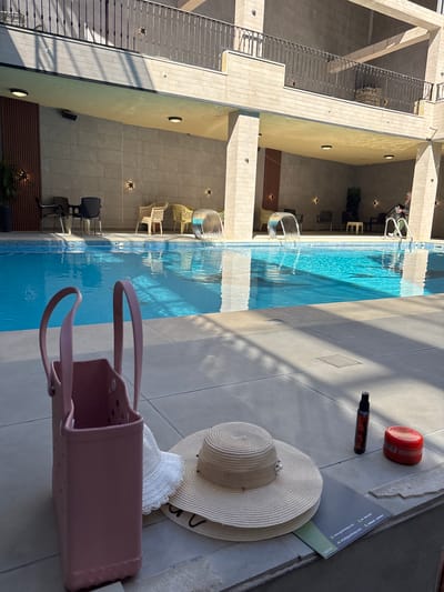 The image depicts an outdoor swimming pool area, likely at a hotel or resort, in Ayn al Basha, Jordan. The pool is filled with clear blue water and features cascading waterfalls on one side. Around the pool, there are lounge chairs and tables. In the foreground, on the tiled pool deck, a pink tote bag, a white sun hat, and a straw sun hat are placed near a red jar and a dark spray bottle. The architecture suggests a modern building with stone facades and a balcony railing. The lighting indicates it is daytime, with shadows cast by the sun.