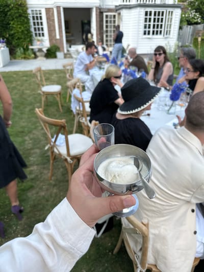A person in a white shirt is holding a small silver cup of ice cream in their left hand and an empty glass in their right hand. The scene is outdoors, likely a garden or lawn. There are several other people seated at tables, some of whom are facing away from the camera. There are flowers on the tables. The setting appears to be a wedding or other celebratory event, with white chairs and tables in the background. A large building is visible in the background. This image does not contain enough evidence to confirm it was taken in the city of Cambridge, United Kingdom.