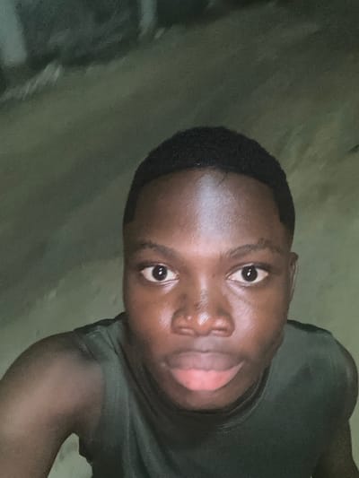 A close-up portrait of a young Black man. He has short, dark hair and shiny skin. His eyes are open, and his mouth is slightly open, with his tongue visible. He is wearing a dark green shirt. The image appears to be taken indoors, with an unclear background. The photo is rotated slightly counter-clockwise. The lighting is dim, casting shadows on parts of his face and shirt.
