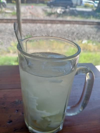 A close-up, eye-level shot shows a clear glass mug filled with a pale yellow, cloudy liquid and ice. A silver spoon rests in the mug, its handle extending out of the top. The mug sits on a brown wooden table. In the blurred background, a railroad track runs horizontally, with ballast stones and scattered green foliage. Beyond the tracks, the blurry shapes of vehicles and possibly buildings are visible, suggesting an outdoor or semi-outdoor setting, likely in Sruni, Indonesia.
