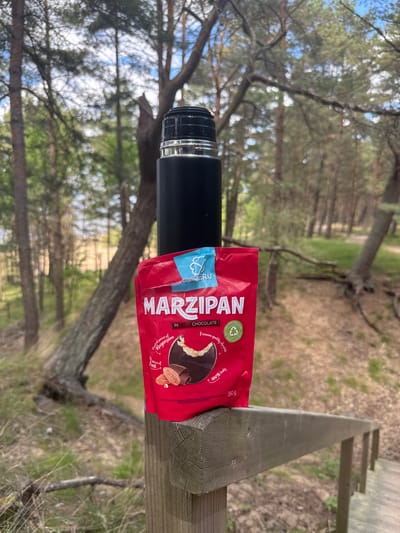 The image captures an outdoor scene in Saulkrasti, Latvia, featuring a black thermos bottle and a red package of marzipan candy resting on a weathered wooden beam, possibly a railing or part of a boardwalk. In the background, a blurred forest or wooded area is visible, dominated by pine trees with green foliage and patches of bright sky filtering through the canopy. The ground beneath the trees appears to be covered with dry leaves and some sparse grass.
The red package prominently displays "MARZIPAN IN DARK CHOCOLATE." Additional text on the packaging highlights its qualities: "Premium quality chocolate" and "100% tasty." It also boasts nutritional benefits, stating "Excellent source of Magnesium" and "Iron." A small, partially visible blue label attached to the thermos reads "SERU." The package further notes "ECO FRIENDLY PACKAGING" and "RECYCLABLE ONLY," accompanied by a circular eco-friendly symbol. The weight is indicated as "150 g."
The scene suggests a break or pause during an outdoor activity, such as a hike or a walk through the forest. The presence of the thermos implies a hot or cold beverage accompanying the snack. The bright lighting indicates it is daytime, likely a clear or partly cloudy day. The setting evokes a sense of tranquility and a connection with nature, typical of recreational areas in places like Saulkrasti.