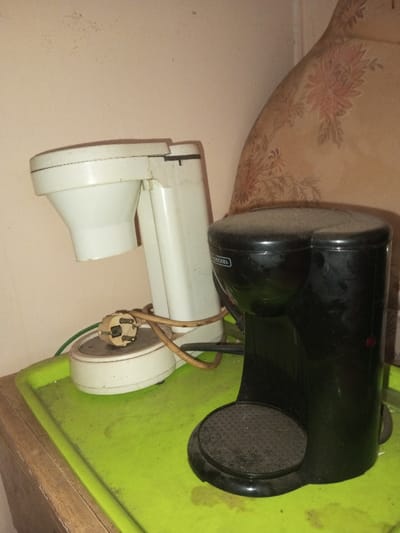 Two dusty coffee makers sit on a green surface, likely a tray, amidst an aged setting. The left coffee maker is white, resembling a vintage model with a circular base and an overhanging filter section. Its power cord is tangled. The right coffee maker is black, a more compact, modern design. Both appear neglected, covered in a layer of dust, indicating a period of disuse. A patterned fabric, possibly a cushion, is behind the objects and the wall is in a pastel color.