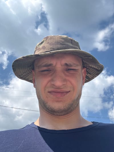 A low-angle, close-up, selfie-style photograph captures a light-skinned male with short, dark hair, a trimmed beard, and a camouflage bucket hat. He is wearing a dark blue t-shirt. The man's face is contorted in a grimace, with his eyebrows furrowed and his mouth slightly open. He is outdoors, with a bright blue sky dotted with white clouds in the background. A few thin, dark wires are visible stretched across the sky, suggesting the presence of utility lines. There is no visible indication of the specific location being Kamensk-Uralsky, Russia; the scene is generic and could be anywhere outdoors on a sunny day with power lines.
