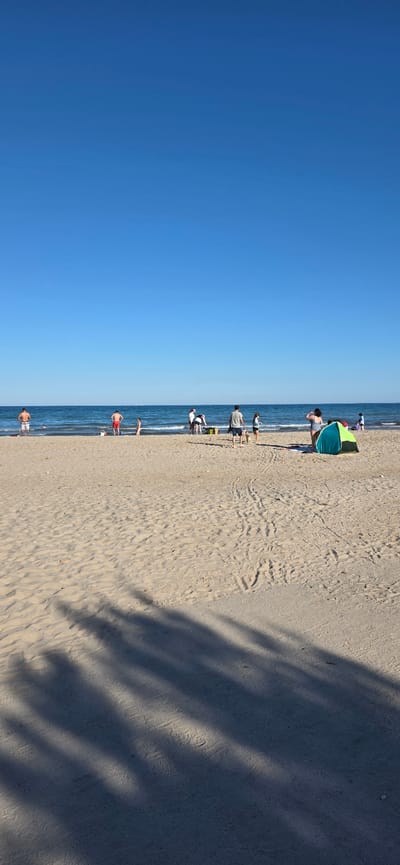 This is an outdoor photograph of a beach in Canet d'en Berenguer, Spain, taken on a sunny day with a clear blue sky. The photograph is oriented vertically, with the sky occupying the left half of the frame and the beach and sea on the right. The beach is sandy, with some areas showing patterns of footprints and wave marks. The sea is a deep blue, and there are several people in the water or near the shoreline. Some people are standing or walking along the shore, while others appear to be swimming or playing in the water. There is a green and blue beach tent set up on the sand. The right part of the photo has shadows from the trees.