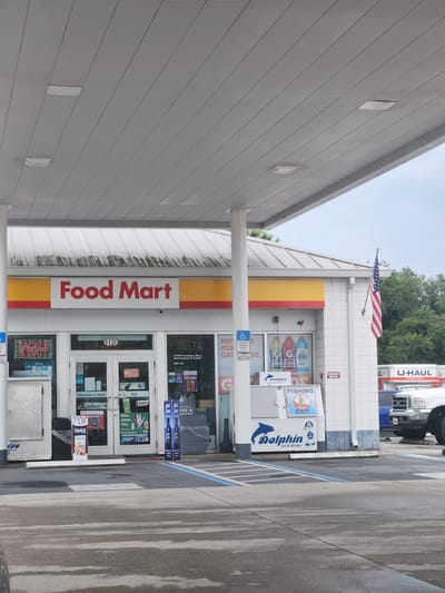 This is a wide outdoor, eye-level shot of a Shell gas station and convenience store called "Food Mart" in Merritt Island, Florida. The building has a white exterior with red and yellow accents. The store features glass double doors that lead inside. A sign above the doors reads "Food Mart" in large red letters. To the left of the entrance, a sign advertises "TACKLE & BAIT". To the right, a sign for "DELTA 3120 N Courtenay Pkwy Merritt Island, FL 32953" is visible. An American flag flies from a pole on the right side of the building. A U-Haul truck is parked to the right of the store. The ground is paved concrete, with blue lines marking parking spaces and a gas pump visible on the left. The sky is overcast.