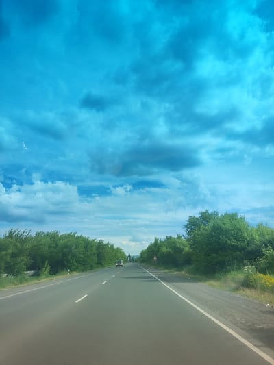The image depicts a long, empty asphalt road stretching into the distance, flanked by lush green trees on both sides. The sky above is a vibrant, deep blue with scattered white and grey clouds. The perspective suggests the photo was taken from a moving vehicle, as indicated by the road markings and the blur of the trees. A white car is visible on the road ahead, appearing to be the only other vehicle in sight. The overall scene evokes a sense of travel and openness, common in rural or semi-rural settings. The location is identified as Azatan, Armenia.