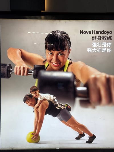 This is a photograph of an advertisement for "Nove Handoyo" fitness training. The advertisement features two women and a child. In the foreground, a woman is performing a plank position with a child on her back, her hands resting on a yellow exercise ball. In the background, another woman is lifting dumbbells. Text on the right side of the advertisement includes "Nove Handoyo" and Chinese characters that translate to "Fitness Training" and "Strong is you, Powerful is you." The advertisement appears to be displayed in an indoor setting, likely a public space such as a shopping mall or transit station in the Xuhui District of China, given the presence of Chinese text and the context of the request.