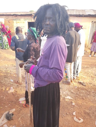 A person with dark skin and wild, dark hair stands in the foreground, wearing a purple long-sleeve shirt and a long black skirt. They are holding a rope. Behind them, a crowd of people gathers outside a building with the words "Bideenu Orphanage and Saloon" visible. The ground is dirt-covered and littered with debris. Other individuals in the background wear colorful clothing and casual attire.