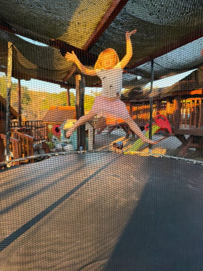 The image captures a view through a black safety net, likely enclosing an outdoor play area. In the foreground, a young child with blonde hair is visible from the chest up, their body bent at the waist as if playing or moving. They are wearing a white t-shirt and a pink skirt. Their right arm is extended outwards, and their left arm is bent, with their hand resting on their hip. The child appears to be in motion, possibly jumping or playing.

Further in the background, through the net and illuminated by warm, golden light, are other figures and structures. One person appears to be wearing a pink shirt. There are wooden structures with horizontal planks, suggesting a playframe or climbing apparatus. Brightly colored objects, including red and yellow, are visible, hinting at toys or play equipment. The lighting suggests it is either sunrise or sunset, casting long shadows. The overall scene appears to be an outdoor recreational area, possibly a playground or a trampoline park, given the netting. The backdrop shows hints of natural scenery with greenery and possibly hills or mountains, consistent with a location like Zagora, Montenegro. The mood seems lively and playful, with the children engaged in activity.