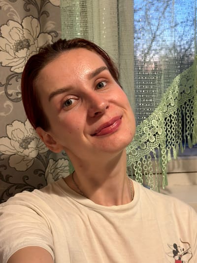 A woman with fair skin, auburn hair, and light eyes poses for a selfie-style photograph. She wears a white t-shirt with a graphic design visible on the upper right side. A thin silver chain is visible around her neck. Her facial expression is a slight smile. The background includes a window with a light green, lace-like curtain partially covering it. A gray and white floral patterned fabric is visible beneath her. The location cannot be determined based on the image. No details related to Заболотово, Russia are visible.