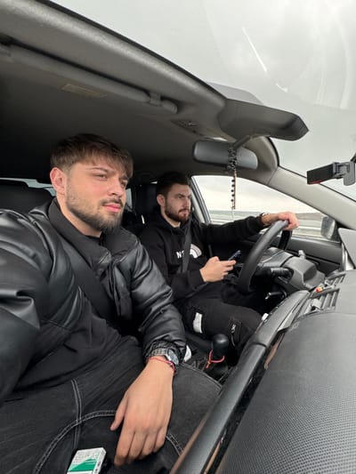 The image depicts the interior of a vehicle traveling on a road in Mlada Gvardia, Bulgaria, under an overcast sky. Two adult males occupy the front seats. In the foreground, the passenger, a male with dark brown hair and a short beard, is seated on the right, wearing a black puffer jacket, a black shirt, and dark jeans with white stitching. A silver wristwatch and a red and white string bracelet are on his left wrist. A pack of "DELIA FLAME" cigarettes rests on his lap. He is buckled into a seatbelt and looking slightly to his right, away from the camera. The driver, also a male with dark brown hair and a short beard, is seated on the left, wearing a black hoodie with a partially visible "THE NORTH FACE" logo and dark cargo pants. His left hand grips the steering wheel, while his right hand holds a dark-colored smartphone. A silver watch with a brown strap is on his left wrist. A black rosary hangs from the rearview mirror. A phone mount is affixed to the windshield on the passenger side. The vehicle's interior features a dark gray dashboard with air vents and a manual gear shift lever adorned with red and yellow string. The road outside features guardrails.