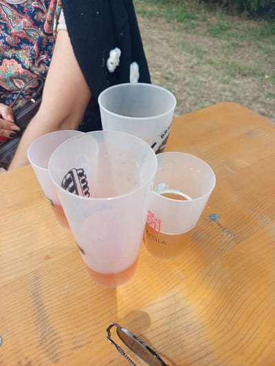 Four translucent plastic cups containing beverage are arranged on a light brown wooden table outdoors. One cup, closer to the foreground on the right, contains a golden-colored liquid with a frothy head, and has "ZKOŁA" printed on it in red. Another cup, to its left, is partially filled with a pinkish liquid and has a black and white illustration of a microphone inside. A third cup, further to the left, also contains a pinkish liquid. The fourth cup, on the far right and behind the others, is empty and has the word "BRA" visible on its side. The background shows green grass and foliage, indicating an outdoor setting. In the upper left, a person's arm wearing a colorful patterned shirt and a black cardigan is visible, along with a dark handbag. The lighting suggests it is daytime.