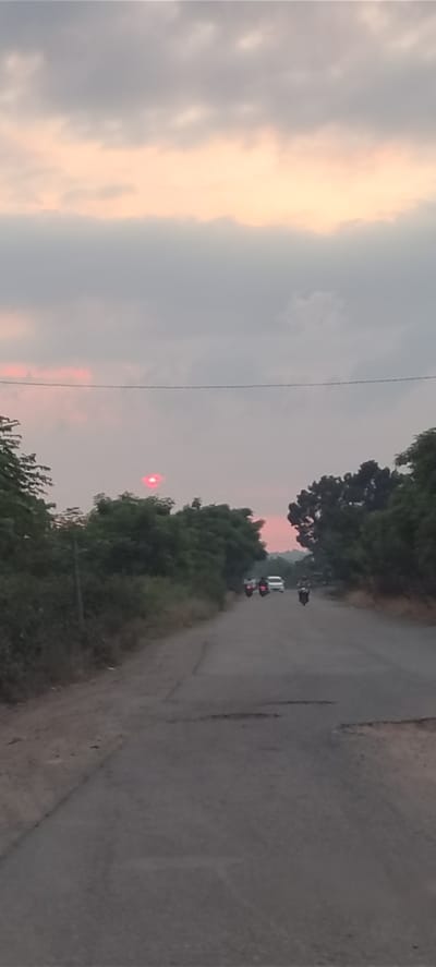 The image depicts a rural road scene at dusk in Blangmajron, Indonesia. A paved road, showing signs of wear and tear with visible potholes, stretches into the distance. Flanked by lush green trees on either side, the road appears to be a typical pathway in a village setting. The sky is overcast with soft pink and orange hues indicating the setting sun. A few motorcycles are visible on the road, with riders in motion, suggesting daily commute or travel. A white car is also present further down the road. In the sky, a bright red circular light is visible, likely a reflection or a distant light source, contributing to the atmospheric quality of the scene.
