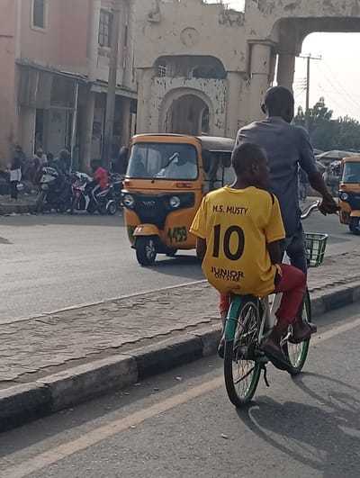 The image presents a daytime street scene in Kofar Mazugal, Nigeria, under clear skies. In the foreground, an adult male cycles a green bicycle, carrying a young boy as a passenger behind him on the rear rack. The boy wears a bright yellow t-shirt featuring the text "M.S. MUSTY" above the large number "10", with "JUNIOR CITY STAR" printed below. He is also wearing red trousers and flip-flops. The adult is dressed in a grey long-sleeved top. Their elongated shadows are cast on the asphalt road to their right, suggesting a morning or late afternoon sun.
To the left of the cyclists, a yellow auto-rickshaw, locally known as a Keke Napep, is visible, bearing the number "4459" on its front. Another auto-rickshaw is partially visible further down the road on the right. In the background, a busy street unfolds with several motorcycles parked or moving, and pedestrians can be seen on the pavement and near the buildings. The environment includes older buildings, notably a large, light-colored structure with prominent archways and some signs of wear, adding to the local character of the scene. A raised sidewalk with a dark curb runs alongside the road. The image captures the everyday activity and diverse transportation methods characteristic of an urban setting in Nigeria.