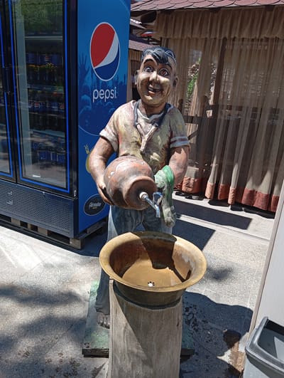 The image shows a statue of a man holding a jug with a tap over a basin. This statue is located in Chinarkent, Uzbekistan. The statue is made of painted concrete. The man has black hair, reddish-brown skin, and wears a light-colored shirt and jeans. He is smiling, and his eyes are wide open. The jug he holds is brown. The basin is brass. Behind the statue is a Pepsi refrigerator full of bottles, and to the right is a canopy with brown curtains. The ground is paved with gray stone tiles.