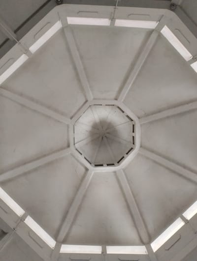 The image shows a low-angle view of a white octagonal ceiling with radiating beams. There are rectangular light fixtures positioned between the beams on the outer edges of the octagon. The central part of the ceiling has a circular shadow cast upon it, suggesting a light source from above or an object within the structure. The overall impression is of an interior architectural space, possibly a pavilion or a room with a domed or multi-faceted ceiling. The specific location in Tanjungsari, Indonesia, is not visually indicated in the image itself.
