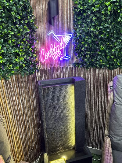 The image depicts an outdoor patio area, possibly in a residential or commercial setting, in Belfast City, United Kingdom. The focal point is a vibrant pink and blue neon sign that reads "Cocktail Bar" with an illustration of a cocktail glass and straw. This sign is mounted on a backdrop of vertical bamboo poles, partially covered by artificial green foliage on either side. In the foreground, a modern, dark-colored water fountain with a textured surface cascades water, illuminated by a yellow light. To the right of the fountain, a portion of a grey upholstered armchair or sofa is visible, along with a light-colored wicker chair. The ground appears to be covered in artificial turf. The overall impression is of a stylish and inviting space designed for relaxation and socializing.
