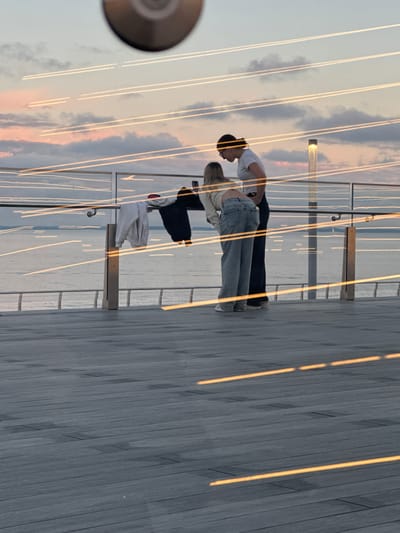 The image shows two people standing on a wooden deck. The person on the left is wearing a white t-shirt and dark pants, with their head bowed. The person on the right is wearing a light-colored top and jeans, also with their head bowed, seemingly in conversation with the first person. There are railings visible behind them, suggesting they are on a balcony, pier, or elevated walkway. The background appears to be a body of water with a cloudy sky, with hints of pink and orange suggesting it might be sunset or sunrise. The wooden deck has a reflective surface, creating streaks of light that are visible across the image. There is also a circular object partially visible on the left side of the frame, which could be a lens or a decorative element. The overall mood is serene and intimate.