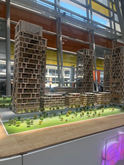 This media file shows a detailed architectural model of a futuristic city with modern buildings and lush green spaces. The buildings are characterized by their glass facades and intricate grid-like structures, suggesting a contemporary architectural style. Several miniature trees are strategically placed throughout the model, adding a touch of realism to the urban landscape. The scene is illuminated by bright, natural light, indicating it's daytime.
The camera angle is high and angled, offering a broad view of the model. In the background, a large-scale, glowing, purple sign that appears to say "5G" is visible, suggesting the model might be part of an exhibition or presentation related to telecommunications technology. The overall setting appears to be indoors, possibly a museum, exhibition hall, or a corporate display area, given the clean, well-lit environment and the presence of the large sign. The scene is tranquil and static, depicting a constructed environment rather than an active event. There are no discernible people actively engaged in any activity within the model itself, but there are blurred figures visible through the glass facades of some of the buildings and in the background, suggesting people are present in the surrounding area. The location context provided is Parakar, Armenia.