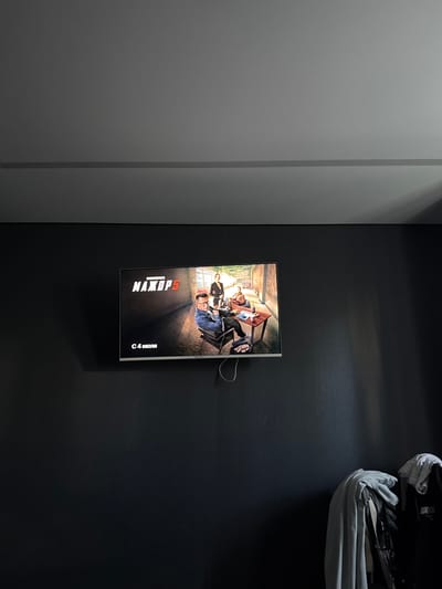 The image shows a modern interior space, likely a living room or entertainment area, with a dark, textured wall. A flat-screen television is mounted on the wall, displaying a movie poster or title screen. The screen features a title in large, bold, stylized Russian letters that read "МАХОР 5", with smaller text above it that appears to be "БОГАТИР". Below the title, text reads "С 4 ИЮЛЯ", indicating a release date of July 4th. The screen also shows an image of several people, possibly actors, seated around a table in an outdoor or semi-outdoor setting, with a hint of greenery in the background.
In the upper right corner of the image, a large, light-colored bundle of fabric, possibly a blanket or duvet, is draped over a surface, partially obscuring a black object, which might be a bag or backpack. The lighting in the room suggests it is daytime, with light casting shadows on the wall from an unseen source, possibly a window or lamp. The overall mood is contemporary and minimalist, with the television and its display serving as the focal point. The location context provided is Velikiye Luki, Russia, which is consistent with the Russian text displayed on the screen.