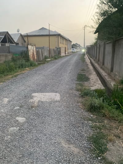 This image depicts a gravel road in a residential area in Боғистон, Tajikistan, likely taken during the late afternoon or early evening. The road is flanked by a concrete drainage ditch and overgrown grass and vegetation on the left, and houses and buildings on the right. The houses appear to be of varying architectural styles, some with yellow facades and dark roofs, and others with more modern corrugated metal roofing. There are several utility poles visible along the road, with power lines stretching across the hazy sky. The atmosphere is slightly overcast, suggesting possible dust or pollution in the air. The road is empty, with no people or vehicles visible, giving a sense of quiet solitude to the scene. The gravel surface of the road is rough, with some patches of lighter-colored stones and a few wet spots, perhaps from recent rain or irrigation. The overall impression is of a typical, somewhat unkempt, semi-rural or suburban setting.