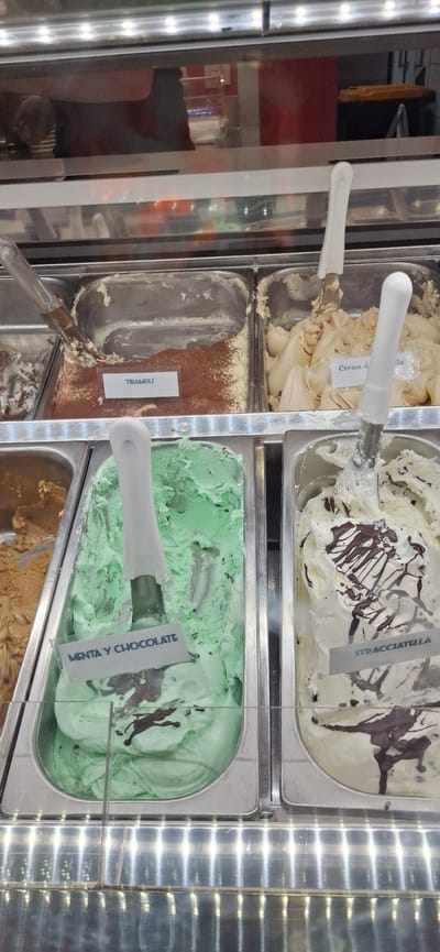 The image shows a refrigerated display case containing several flavors of gelato in a shop likely located in Santa Cruz de Tenerife, Spain. The gelato flavors visible include "Crema d", "Stracciatella", "Tiramisu", and "Menta y Chocolate." Each flavor is in a rectangular metal container with a labeled tag. Scoops are present in the containers, indicating that the gelato is ready to serve. The overall environment suggests a typical gelato shop setting.