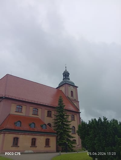 An outdoor, eye-level shot captures a historic-looking building, likely a church or monastery, under a heavily overcast, dull gray sky in Kiełczów, Poland. The main structure features a prominent clock tower with a belfry topped by a cross, indicating its religious nature. The building has reddish-brown tiled roofs and light peach or beige-colored walls. Multiple rectangular windows with dark frames are visible across its facade.

To the left, a smaller, attached section of the building, also with a red tiled roof, includes three distinct arched dormer windows and several rectangular windows on its lower level. Lush green grass occupies the foreground and extends to the right, where a dense cluster of dark green coniferous trees partially obscures the building. Another tall, conical coniferous tree stands directly in front of the main building, partially obscuring the lower part of the tower. A dark, indistinct figure can be faintly seen standing at the base of this large tree. A paved path or courtyard is visible near the bottom of the image, with small modern bollards on the grass.

The weather appears gloomy and overcast, with no sunlight visible, characteristic of a heavily clouded day. The time stamp "05.06.2026 13:23" is visible in the bottom right corner, and "POCO C75" is in the bottom left, indicating the camera or device used. The clock on the tower is visible but too indistinct to precisely read the time.