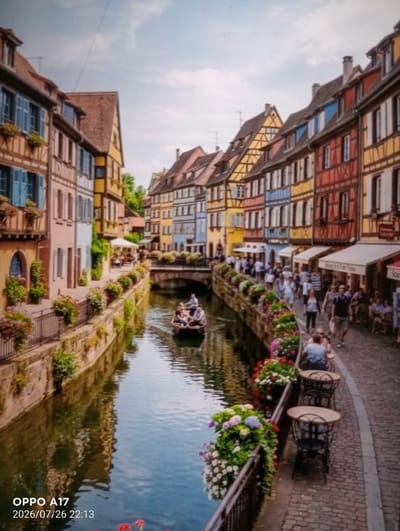 The image depicts a scenic canal lined with colorful, historic buildings. The architecture is characteristic of European towns, with timber-framed facades and steeply pitched roofs. A narrow waterway flows through the center, reflecting the pastel hues of the buildings and the sky. Several people are visible, some strolling along the cobblestone path beside the canal, while others are seated at outdoor tables in what appears to be a cafe. A small boat, carrying a few passengers, is navigating the canal. Lush greenery and vibrant flowers in planters add to the picturesque atmosphere. The sky is partly cloudy, suggesting a pleasant day. The lower left corner of the image displays "OPPO A17" and a date and time stamp, indicating the device and time of capture. This location is likely a tourist destination.