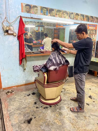This image depicts the interior of a barbershop in Payatieng, Indonesia. A man, presumed to be the barber, is in the process of cutting the hair of a young boy sitting in a barber chair. The barber is using an electric trimmer, and the boy is wearing a barber cape adorned with a pattern of scissors and combs. The floor of the barbershop is littered with hair clippings, indicating recent activity. In the background, a mirror reflects other barbers and customers, as well as a display of framed hairstyles. A red cloth is draped over a rack on the left side of the image, near an electrical outlet and tangled wires. The walls are painted a light blue, and the floor appears to be concrete. The scene is brightly lit, likely by overhead fluorescent lights visible in the mirror's reflection.