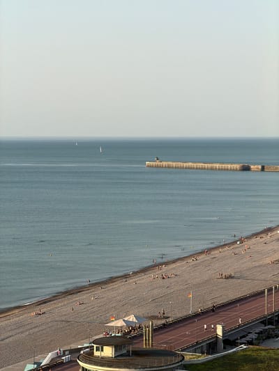 This is an aerial, full-shot, vertical, landscape image taken from a high vantage point, looking down at a beach and the sea. The scene depicts a sunny day at a pebble beach lined with a promenade, where people are engaged in various activities.
In the water, a long, rectangular structure, likely a pier or breakwater, extends from the shore into the sea. There are a few small boats visible in the water, and two even smaller boats or buoys further out. The sea itself is a calm, blue-grey color.
The beach is covered in grey pebbles, and scattered across it are numerous people, some sunbathing, others walking. A paved promenade runs parallel to the beach, with a red track on it, suggesting it may be used for running or cycling. More people are seen on the promenade, and there are lamp posts and a few small structures, including what appears to be a building with a rounded section. Some beach umbrellas are set up, and a flag is visible on a flagpole. The overall atmosphere suggests a leisurely summer day.
The image is well-lit, suggesting it was taken during daylight hours, likely in the late afternoon or early evening given the warm tones on the promenade and the long shadows cast by some of the structures and people. The sky is a light, hazy blue, indicating clear weather. The location is Dieppe, France, as indicated by the context. No visible text is present in the image.