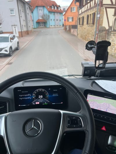 The image displays a forward-facing view from the driver's seat of a Mercedes-Benz vehicle, possibly a truck, in Mühlhausen, Germany. The digital instrument cluster shows a speed of 28 km/h, an odometer reading of 8730.0 km, and an outside temperature of 8°C. The steering wheel is black leather with chrome accents and integrated controls. To the right of the steering wheel, a digital touchscreen displays a navigation map. A suction-cup mounted phone holder or device mount is affixed to the upper right portion of the windshield, below which another black electronic device is present on the dashboard. A paper document is visible on the dashboard to the right.
The vehicle is positioned on a paved road, which appears to be a two-lane street without visible markings, curving slightly to the right and ascending gently. On the left side of the street, a white Hyundai Tucson SUV is parked. Behind it are multiple residential buildings of varying colors, including a light gray structure, a light blue building with a reddish roof, and an orange building. On the right side of the street, an older half-timbered building with stone or stucco siding and a brown-tiled roof is prominent, followed by another orange building. The sky is overcast. No individuals are visible. The scene depicts a quiet street in a European town, consistent with Mühlhausen.