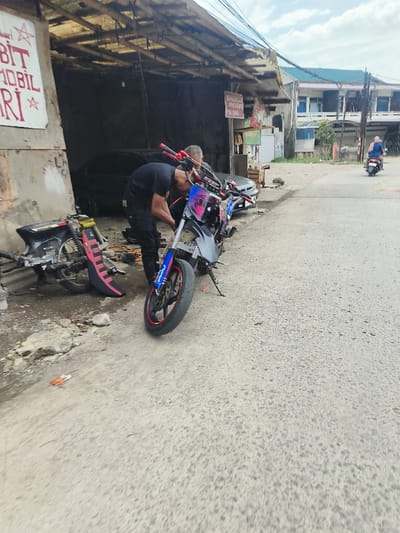 The image captures an outdoor scene, likely a street adjacent to a motorcycle workshop or garage, in what appears to be an urban or semi-urban area in Southeast Asia, possibly Indonesia. The time of day is daylight, indicated by bright natural lighting, with a partly cloudy or overcast sky.

In the foreground, two men are focused on a customized black and blue motorcycle with red accents on the handlebars and wheel rim. The motorcycle is parked on its kickstand. The man in the black t-shirt and black pants is bent over, seemingly working on or inspecting the motorcycle's engine area. Behind him, another man, older with lighter hair and wearing a light-colored shirt, is partially visible, also looking towards the motorcycle. The motorcycle features a distinctive angular headlight area with a graphic resembling an eye. A license plate, partially obscured, reads "B 24...".

To the left of the working men, another motorcycle frame or a heavily stripped-down motorcycle body lies on the ground, predominantly black with a bright pink or red component that looks like a seat or body panel.

Behind the men and the motorcycles is an open-front workshop or garage area. This structure has a simple, somewhat weathered corrugated metal roof. Inside, a dark-colored car, possibly black, is partially visible. The workshop area appears functional and cluttered with various tools and parts, typical of a mechanic's shop.

Several signs are visible on the workshop's structure. On the far left, a large white sign with red text partially reads "BIT," "MOBIL" (Indonesian for "car"), and "ARI," along with a star symbol. A smaller, darker sign, possibly wooden, hangs from a post further inside the workshop. Although difficult to read entirely, it appears to say "BENGKEL MOTOR" (Indonesian for "motorcycle workshop") or similar, given the context.

In the background, across the asphalt road, a person riding a scooter is moving away from the viewer. Further down the street, there are other buildings, including a multi-story structure with a distinctive green roof. Overhead, multiple electrical or communication wires crisscross the sky. The ground in the foreground and midground is asphalt or concrete, showing some wear and minor debris.